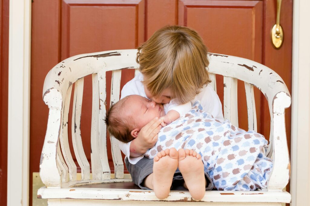 A toddler gently kissing a newborn baby on a white bench