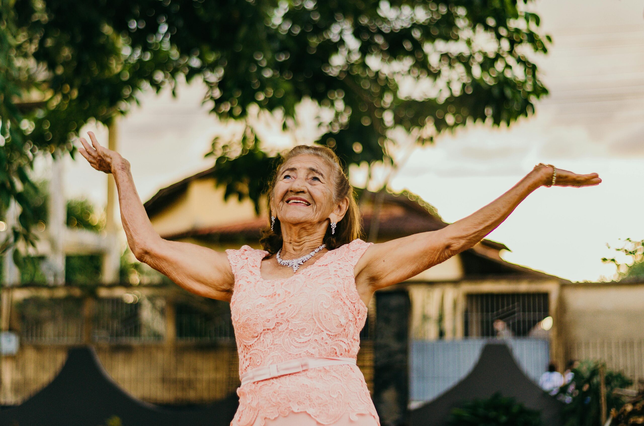 Elderly woman wearing a pink lace dress with arms outstretched, smiling outdoors