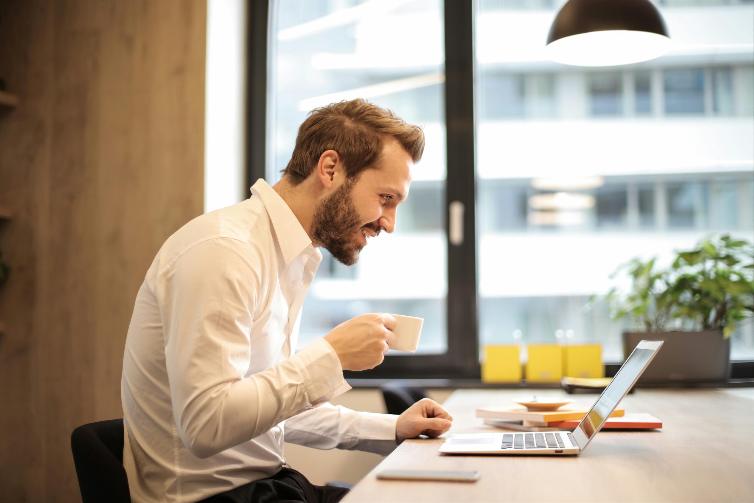 Man in office smiling and holding a coffee cup while working on a laptop